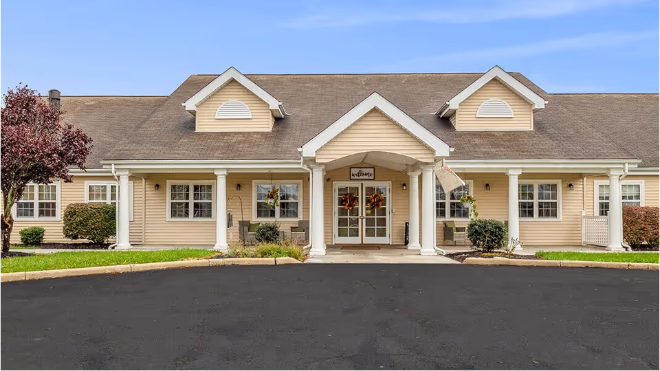 Front entrance of a single-story beige senior living building with white columns, double glass doors and a paved driveway.