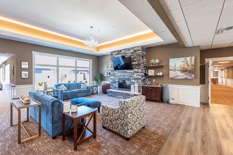 A bright and spacious living room area in a senior living facility featuring two blue sofas, a patterned armchair, a stone fireplace with a mounted flat-screen TV above it, wooden side tables with decorative items, large windows letting in natural light, and a hallway leading to other rooms.