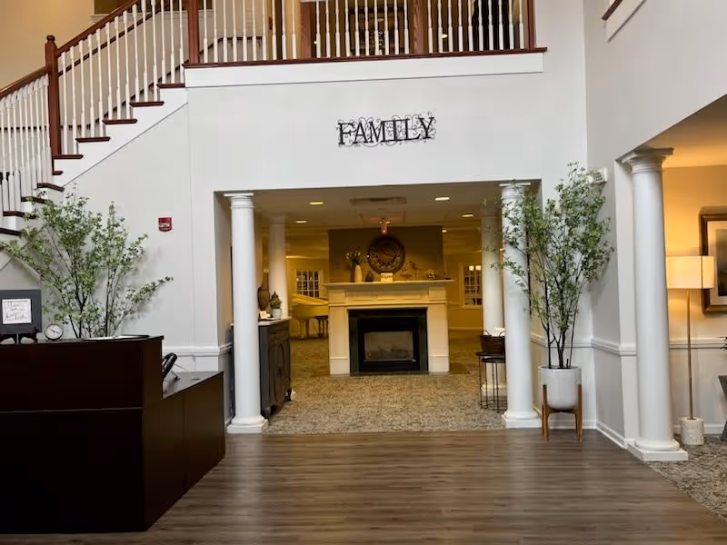 Interior view of a senior living facility lobby area with a wooden floor, two white columns framing a carpeted room with a fireplace, decorative plants on either side, a staircase with wooden handrails on the left, and a wall decoration that reads 'FAMILY' above the fireplace.