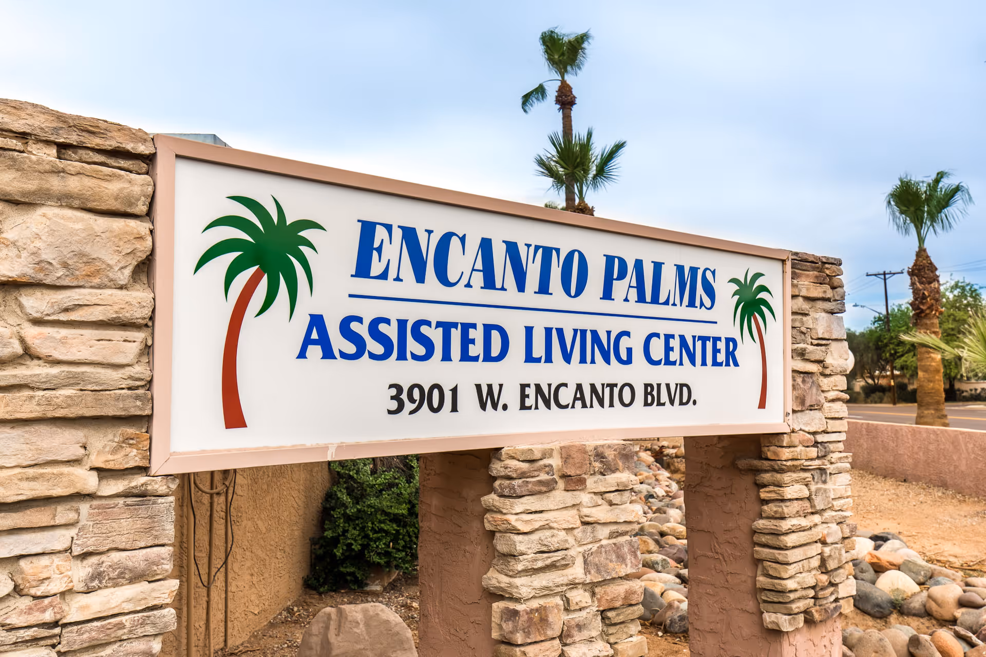 Stone sign for Encanto Palms Assisted Living Center with palm tree graphics on either side, located at 3901 W. Encanto Blvd. with palm trees and a street visible in the background.