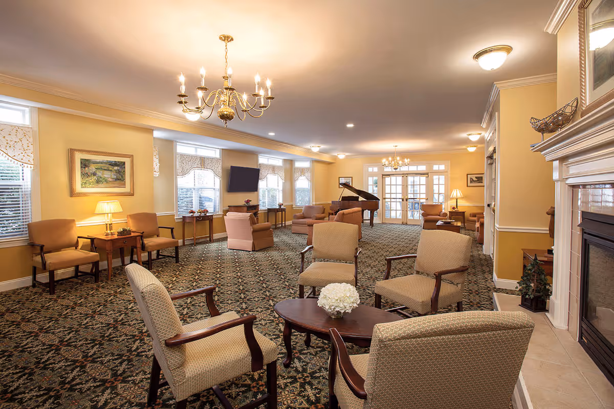 A spacious senior living facility common area with multiple upholstered armchairs arranged around wooden coffee tables. The room features a patterned carpet, yellow walls, several windows with valances, a grand piano in the background, a wall-mounted TV, and a fireplace on the right side. Chandeliers and ceiling lights illuminate the room.