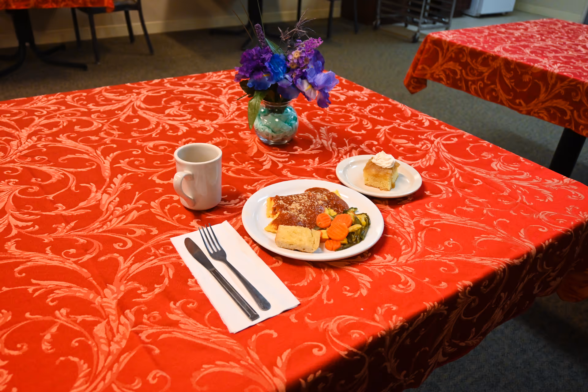 A dining table covered with a red patterned tablecloth. On the table, there is a white plate with a meal consisting of pasta with red sauce, cooked carrots, green beans, and a bread roll. Next to it is a smaller plate with a piece of cake topped with whipped cream. There is also a white coffee mug and a set of silverware (fork and knife) placed on a white napkin. A small vase with purple and blue flowers is also on the table. In the background, other tables with similar red tablecloths and chairs are visible.