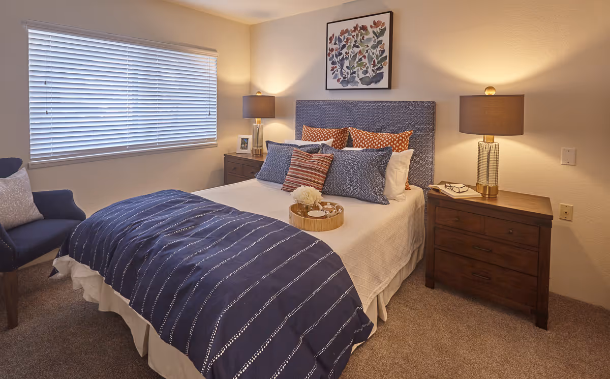 A cozy bedroom with a neatly made bed featuring a blue and white striped blanket and multiple decorative pillows. There are two wooden nightstands on either side of the bed, each with a lamp. A framed floral artwork hangs above the bed, and a window with closed blinds is on the left wall. A blue armchair with a cushion is positioned near the window.