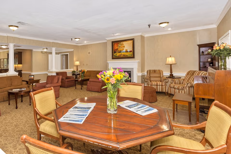 Communal living room with a wooden table and vase of flowers in the foreground, surrounded by sofas, armchairs, a fireplace, and lamps.