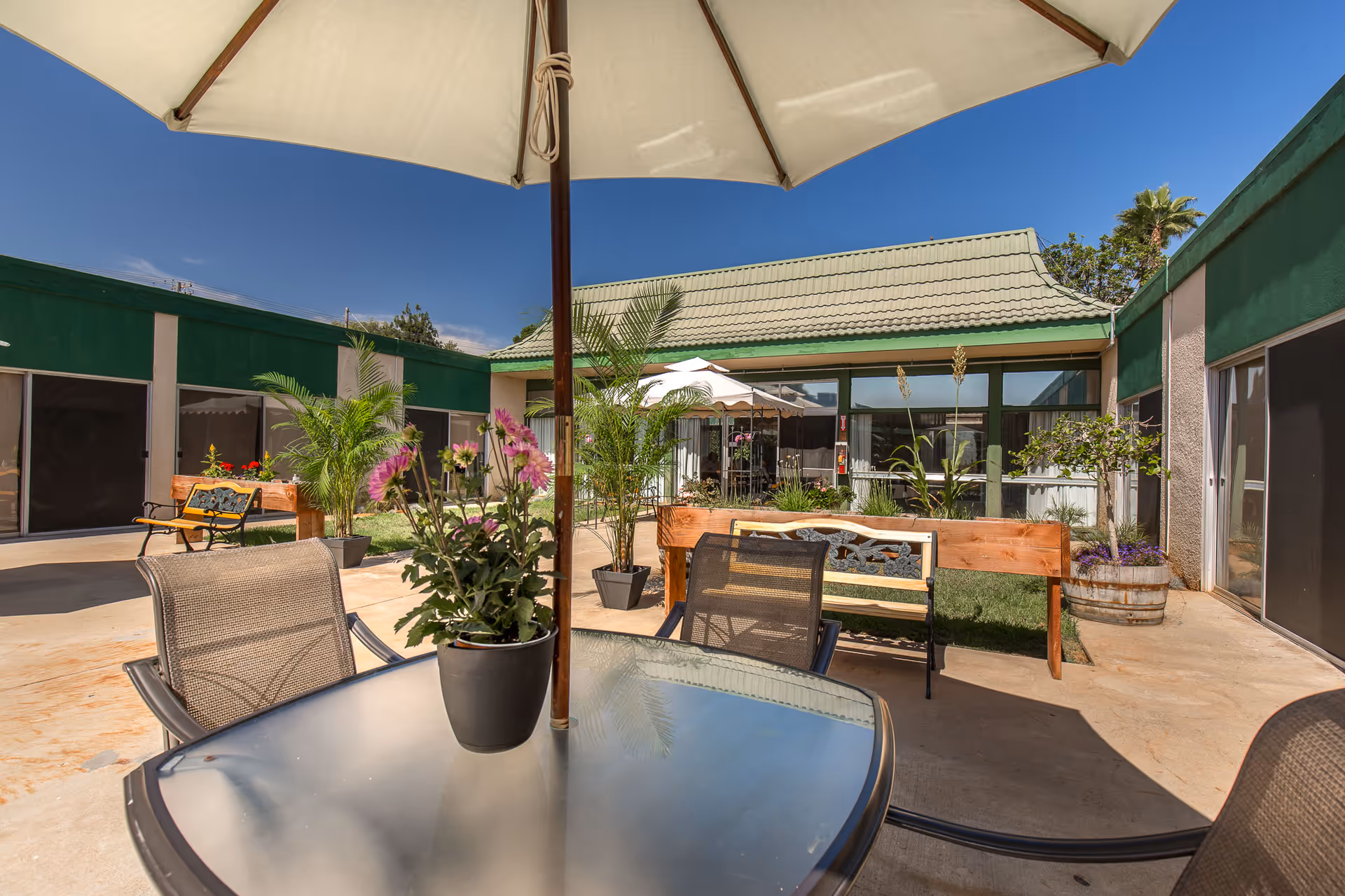 Outdoor patio area at Windsor Gardens Convalescent Center of San Diego with a glass table and chairs under a large umbrella, potted plants, benches, and a building with green trim and a green roof under a clear blue sky.