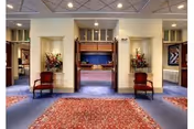 Interior view of a senior living facility lobby or common area with a patterned red carpet, two wooden chairs with red cushions placed symmetrically on either side of a wide doorway, floral arrangements in niches on the walls, and a glimpse of a stage area with blue curtains in the background.