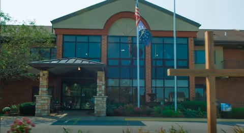 Front exterior view of a brick building with large windows and a covered entrance. Two flagpoles with flags are positioned in front of the building, along with a large wooden cross on the right side. The building is surrounded by greenery and flowers.