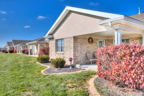 Exterior view of a single-story residential building with beige brick walls and light-colored siding. The building has a covered porch area with chairs and is surrounded by green grass and red-leaved bushes under a clear blue sky.