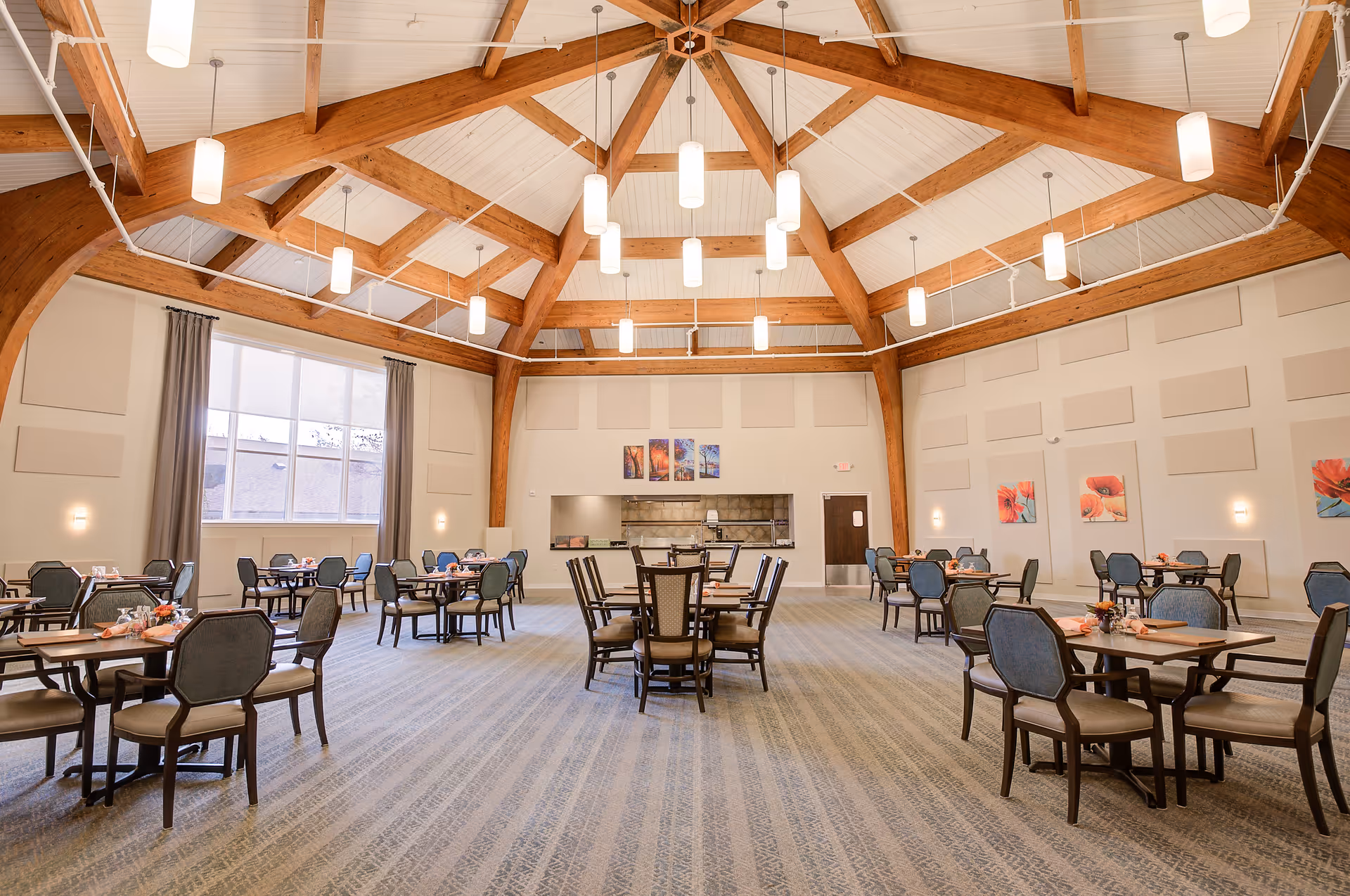 Spacious dining room with multiple tables and chairs arranged neatly. The room features a high vaulted ceiling with exposed wooden beams and hanging cylindrical light fixtures. Large windows with curtains allow natural light to enter. The walls are decorated with colorful floral paintings and acoustic panels. A serving area with a pass-through window is visible at the far end of the room.