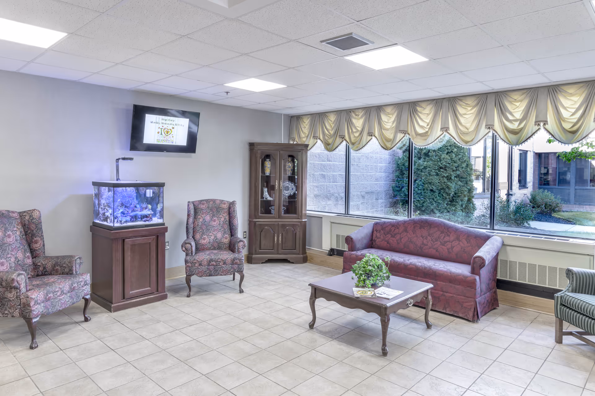 A bright common area with large windows showing outdoor greenery. The room features a maroon sofa, two floral upholstered armchairs, a wooden coffee table with a plant and magazines, a wooden cabinet with decorative items, and a fish tank on a wooden stand. A flat-screen TV is mounted on the wall above the fish tank.