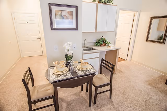 Small dining area with a round glass table set for two with beige dishes and napkins, surrounded by three wooden chairs with beige cushions. Behind the table is a kitchenette with white cabinets, a small sink, and a countertop with a plant. The room has beige carpet, a framed picture on the wall, and a doorway leading to another room.