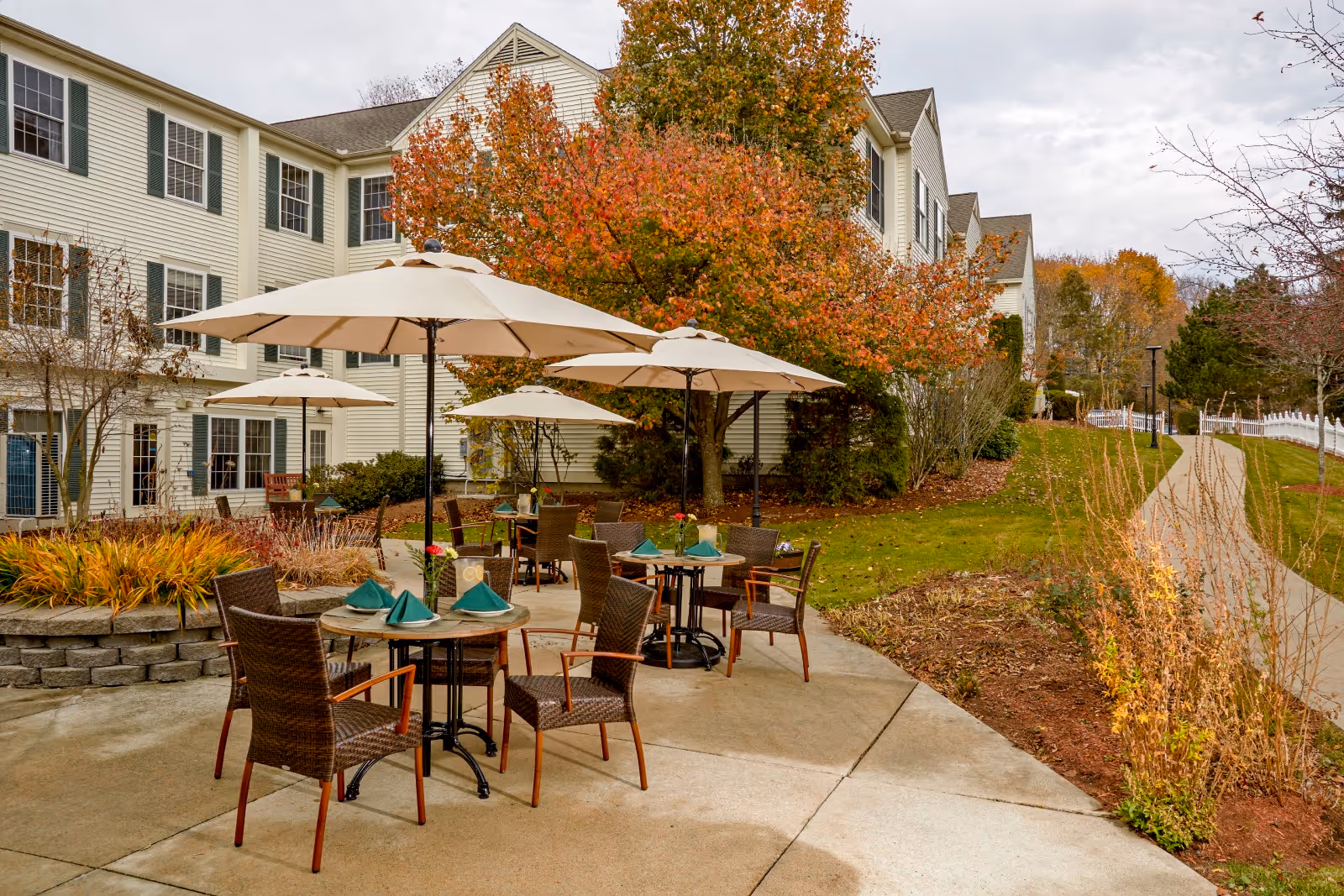 Outdoor patio area at Benchmark at Billerica Crossings with round tables and wicker chairs under large beige umbrellas. The tables are set with green napkins and small flower vases. Surrounding the patio are landscaped plants, a tree with autumn-colored leaves, and a paved walkway leading away from the building.