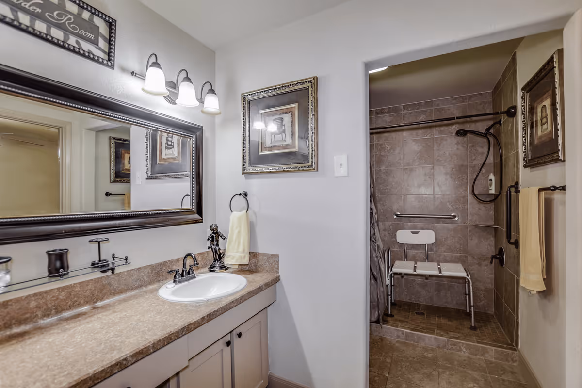 A bathroom with a large mirror above a countertop with a sink. The countertop has a decorative figurine and a towel ring with a yellow towel. The shower area has brown tiles, a shower chair, grab bars, and a handheld showerhead. There are framed pictures on the walls.
