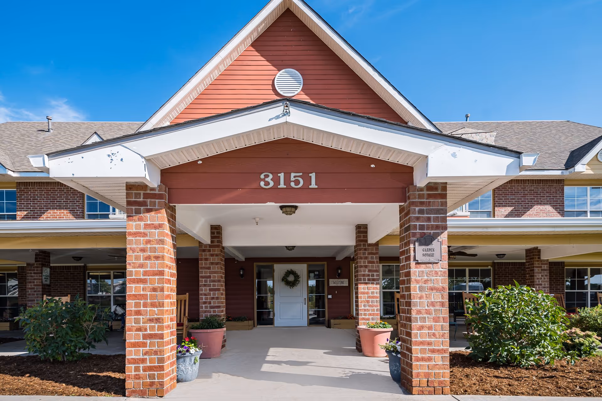 Front entrance of a brick building with a covered porch supported by brick columns, potted plants, and a white door with a wreath. The building number 3151 is displayed above the entrance under a peaked roof.