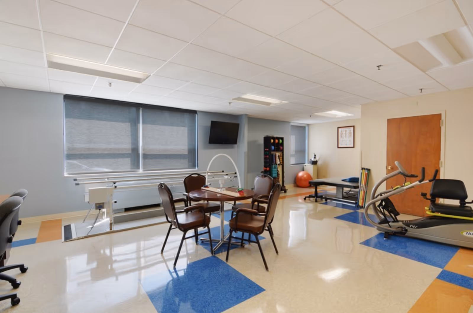 A bright interior therapy room with a table and chairs, exercise equipment, parallel bars, and a TV.