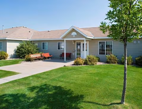 Single-story cottage-style senior living building with a front porch, seating, and a well-kept lawn.
