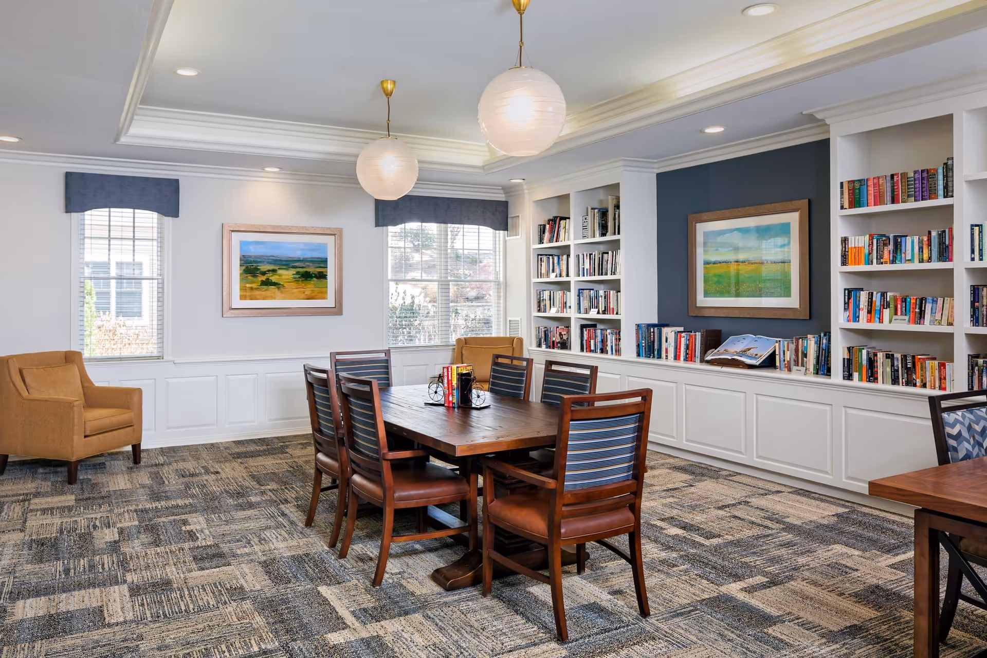 A cozy reading or meeting room with a wooden table surrounded by six chairs, two armchairs near windows with blue valances, built-in white bookshelves filled with books, two framed landscape paintings on the walls, and two round pendant lights hanging from the ceiling.