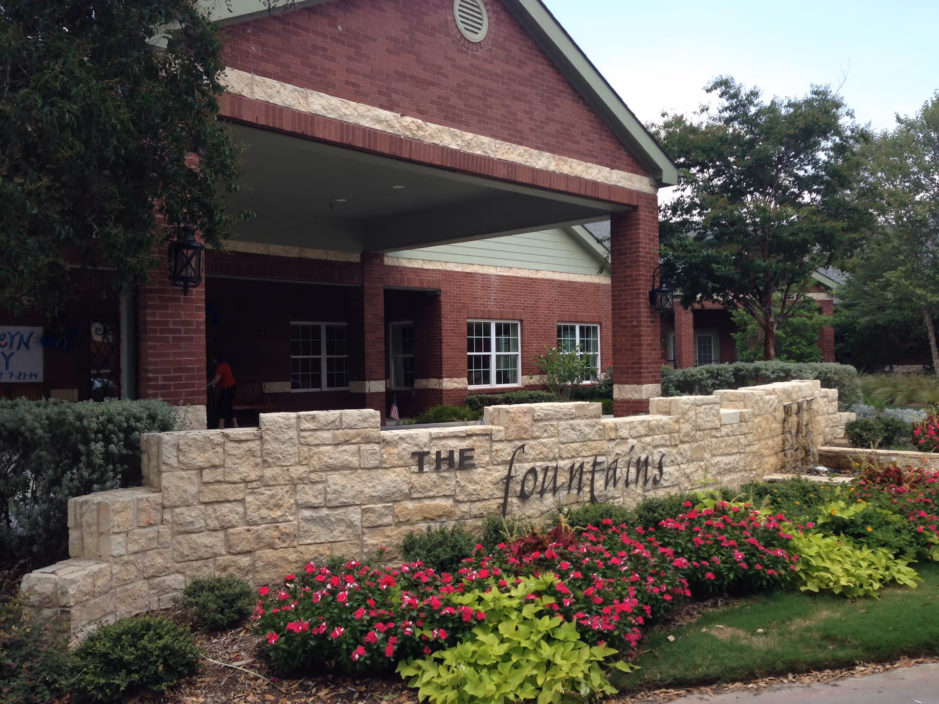 Entrance of a brick building with a covered porch area and a stone sign in front that reads 'The Fountains'. The sign is surrounded by well-maintained landscaping with green shrubs and pink flowers. Trees and additional greenery are visible around the building.