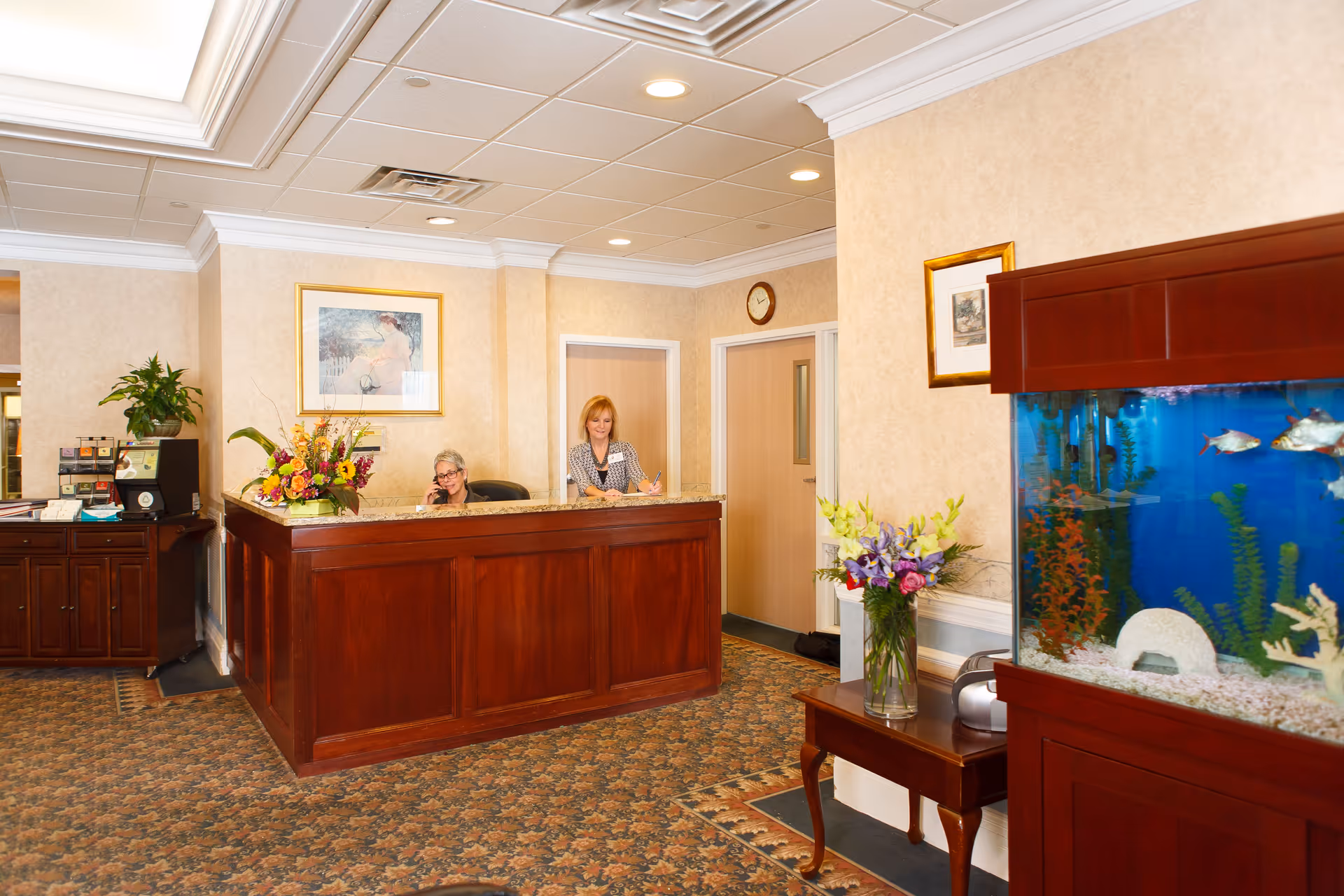 Reception area with a wooden front desk, two staff members behind it, floral arrangements, and a large aquarium.