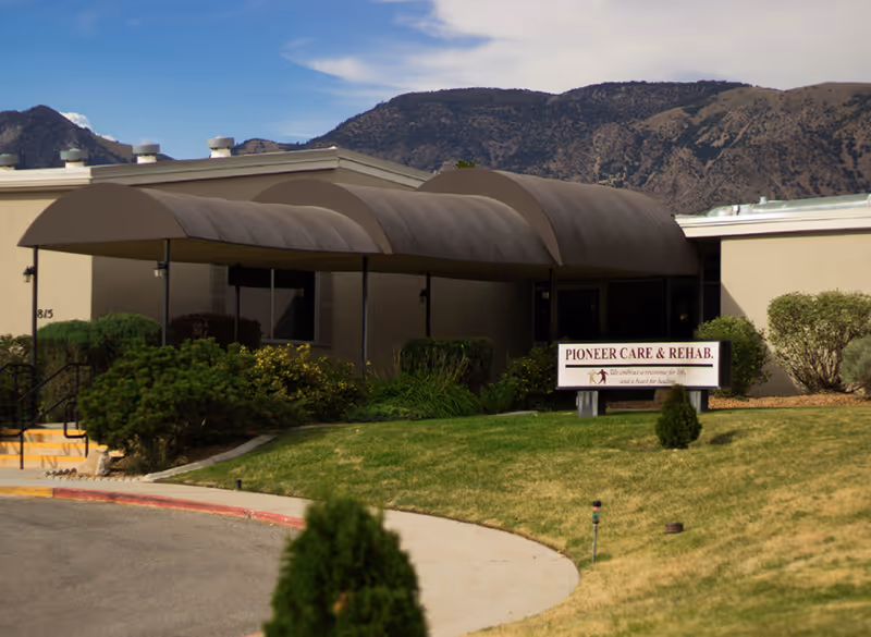 Exterior view of Monument Health Pioneer Trail facility showing a beige building with a curved brown awning over the entrance. There is a sign in front that reads 'Pioneer Care & Rehab' with landscaping including bushes and grass. Mountains are visible in the background under a partly cloudy sky.
