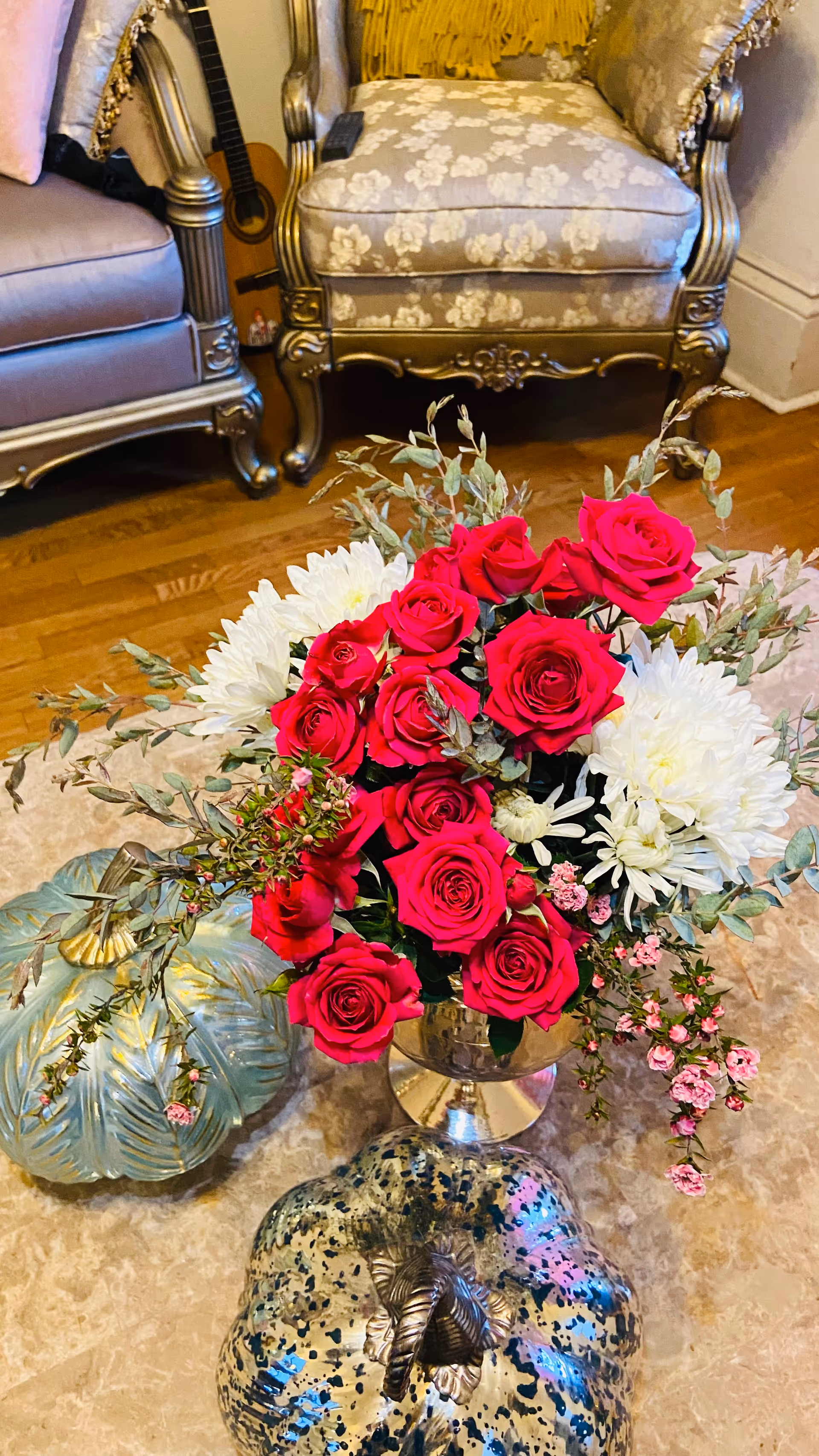 A floral arrangement featuring vibrant red roses, white chrysanthemums, and small pink flowers in a silver vase on a marble table. The table also holds two decorative pumpkins, one blue-green and one speckled silver. In the background, there are two ornate armchairs with floral upholstery and a guitar leaning against the wall.