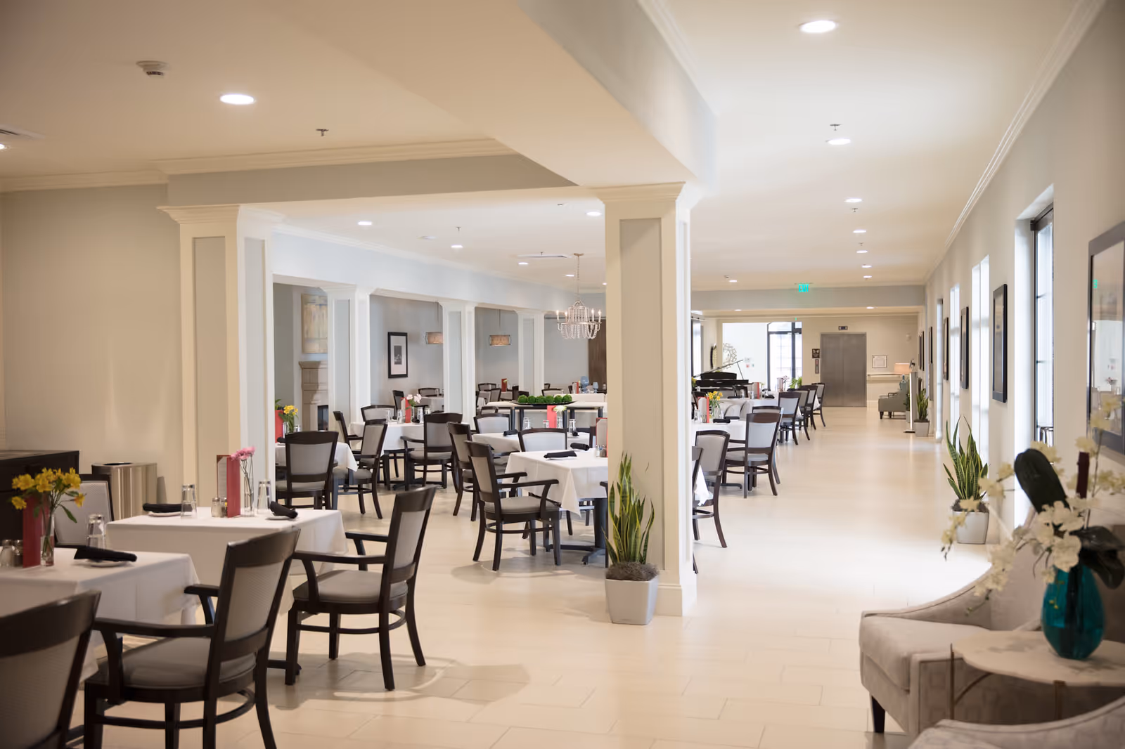 A spacious and bright dining room in a senior living facility with multiple tables covered in white tablecloths, each set with black napkins and glassware. The room features beige walls, large windows on the right side, decorative plants, and comfortable chairs arranged around the tables. The far end of the room has a piano and additional seating, with soft lighting from ceiling fixtures.