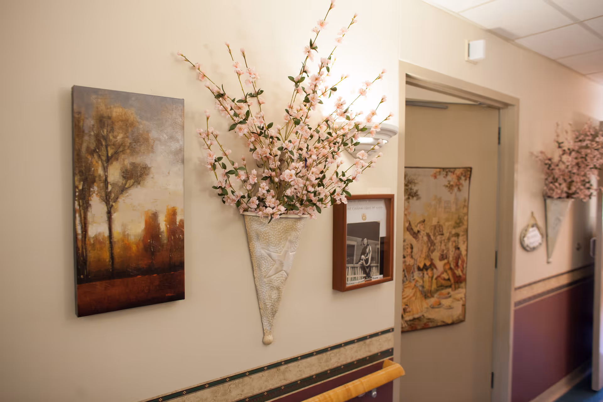A hallway in a senior living facility with beige walls decorated with paintings and wall-mounted vases holding pink artificial flowers. There is a handrail along the wall and a door at the end of the hallway.