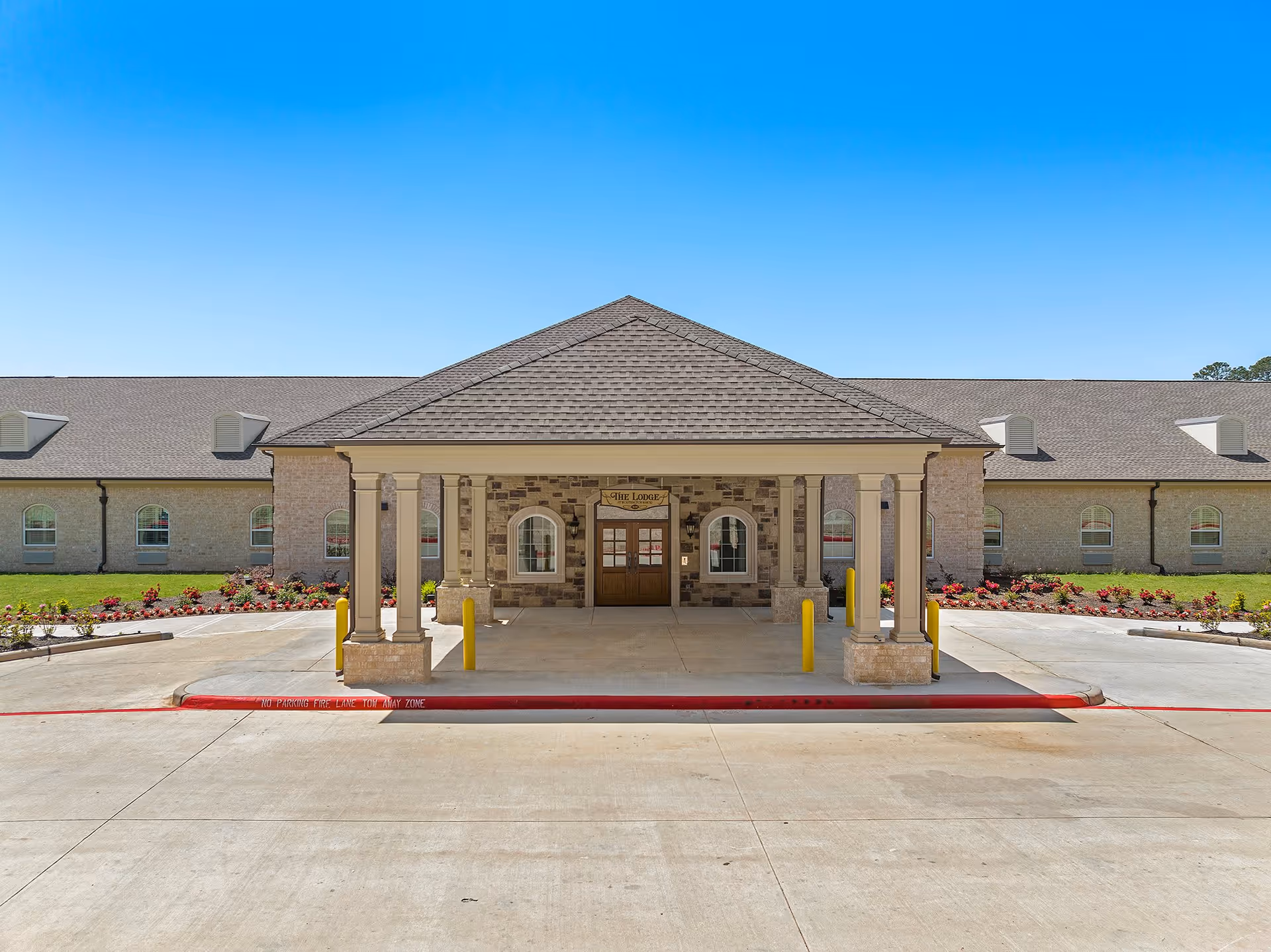 Front exterior view of Worthington Manor showing a single-story building with a covered entrance supported by columns, a sign above the door reading 'The Lodge', landscaped flower beds on either side, and a clear blue sky.