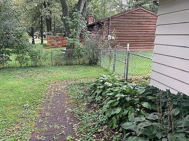 A grassy backyard area with a brick pathway leading to a chain-link gate. There are green plants and bushes along the right side near a white building. In the background, there is a wooden shed and several trees.