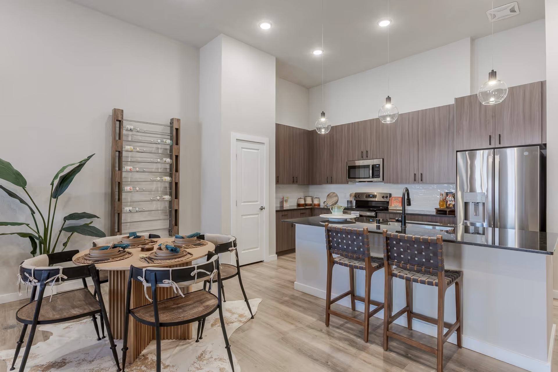 Modern kitchen and dining area with wooden cabinets, stainless steel appliances including a refrigerator and microwave, a black countertop island with two woven bar stools, a round dining table set with plates and napkins, a wine rack against the wall, and pendant lights hanging from the ceiling.