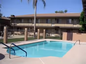 Outdoor swimming pool area with clear blue water, surrounded by a concrete deck and metal handrails. In the background, there is a two-story building with balconies and windows, along with a tall palm tree and some greenery.