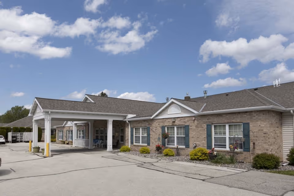 Exterior view of Harbor View Assisted Living facility showing a single-story brick building with a covered entrance, windows with blue shutters, small bushes and flowers along the front, and a clear blue sky with some clouds.