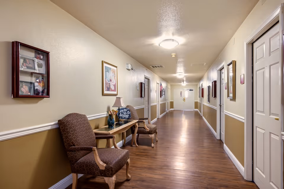 Well-lit interior hallway of a memory care facility with chairs, a side table, framed artwork, and doors lining the corridor.