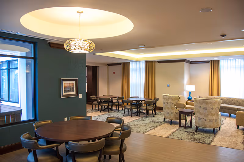 Bright communal lounge with round dining tables, upholstered chairs, and a seating area with armchairs and a sofa.