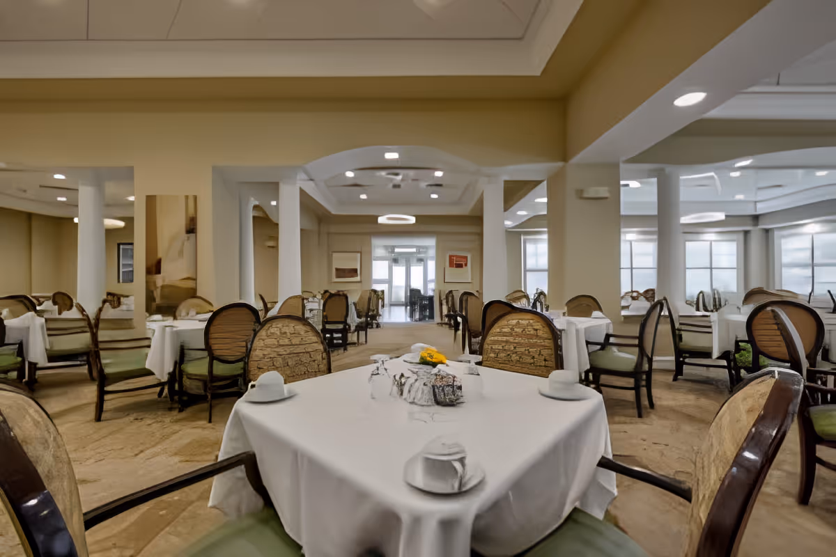 A spacious dining room with multiple tables covered in white tablecloths, each set with cups and glasses. The room features cushioned chairs with wooden frames, beige walls, and large windows allowing natural light. The ceiling has recessed lighting and decorative molding.