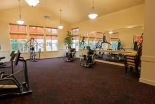 A fitness room with exercise equipment including stationary bikes and weight machines, large windows with blinds, beige walls, carpeted floor, and ceiling lights.