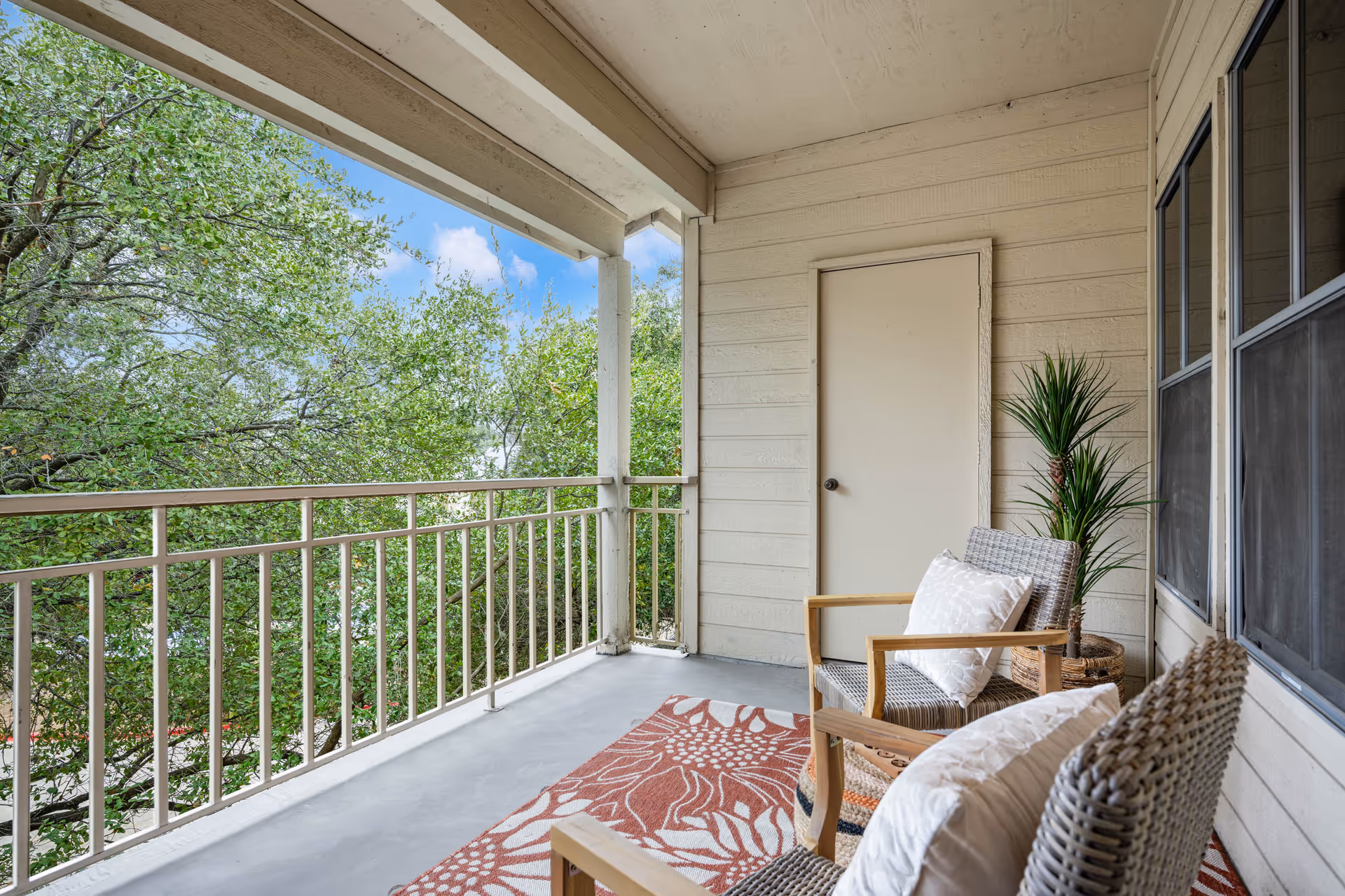 A covered balcony with two wicker chairs with cushions, a patterned rug, a potted plant, and a view of green leafy trees under a blue sky.