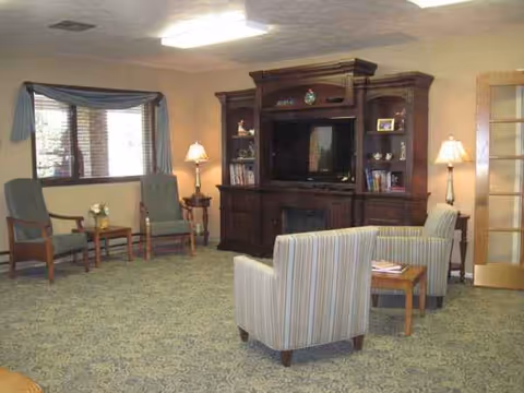 A cozy senior living lounge with striped chairs, lamps, and a large wooden entertainment center.