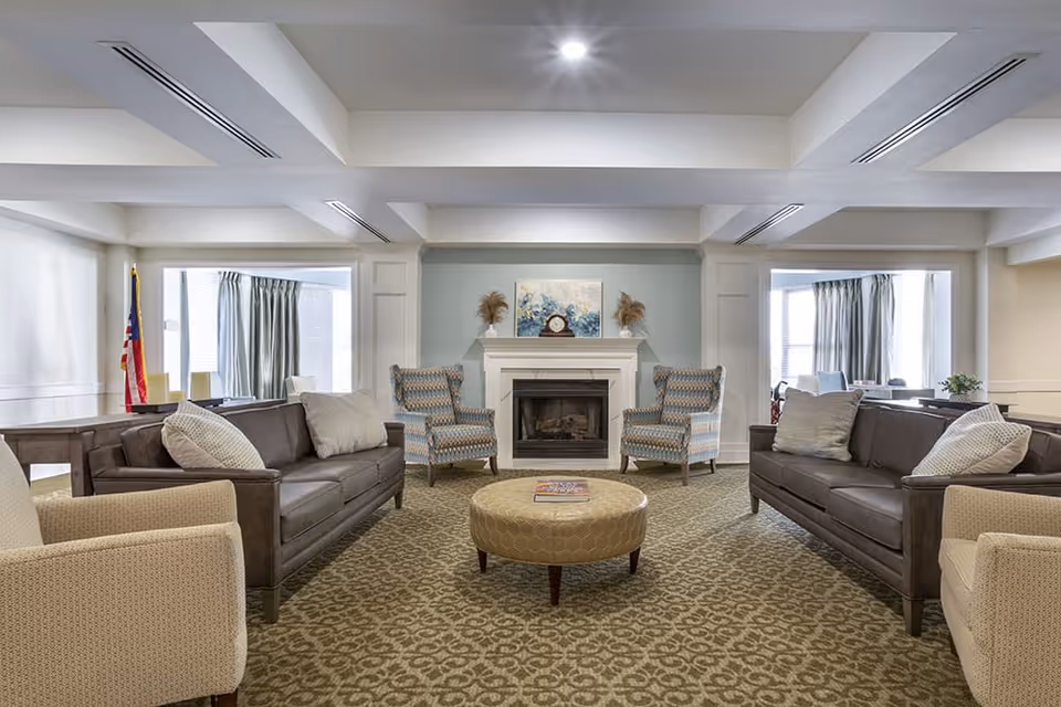 A spacious living room with two brown sofas facing each other, two patterned armchairs near a white fireplace with a clock and decorative items on the mantel. A round ottoman is centered on a patterned carpet. Large windows with curtains allow natural light into the room.