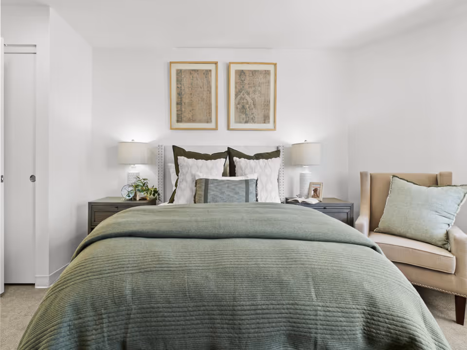 A neatly made bed with a green quilt and multiple pillows in a bright bedroom. There are two framed artworks hanging above the bed. On each side of the bed are dark wooden nightstands with white lamps, a small plant, and a framed photo. To the right of the bed is a beige armchair with a green cushion. The walls are white and the room has a calm, cozy atmosphere.
