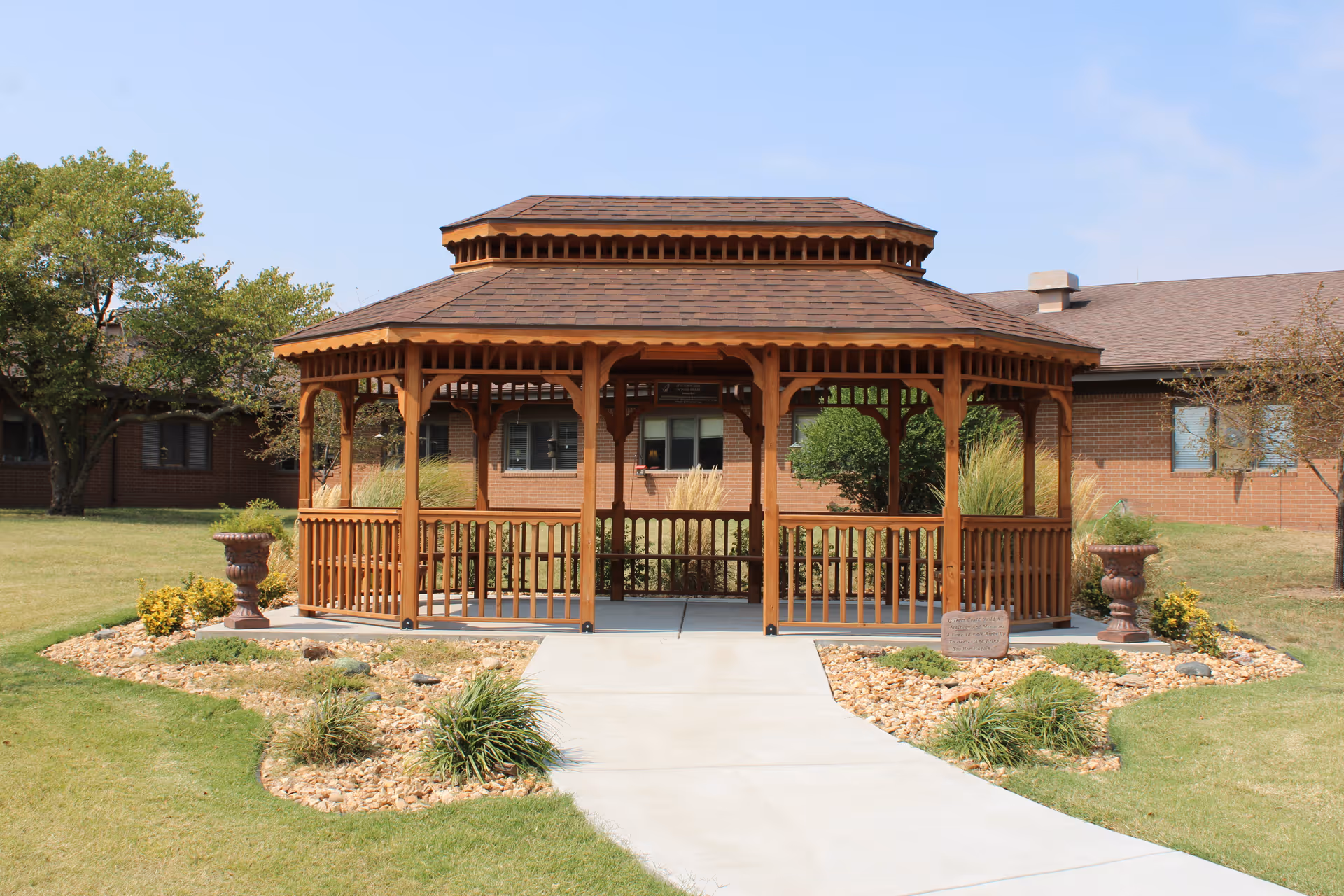 A wooden gazebo stands along a concrete walkway in the landscaped lawn in front of a brick building.