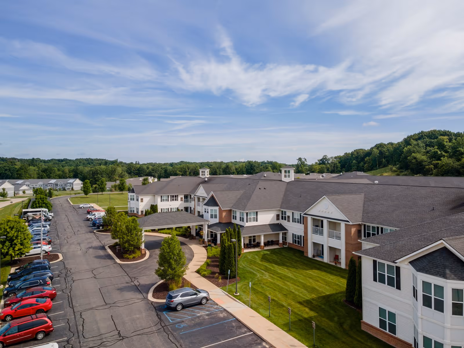 Aerial view of StoryPoint Rockford senior living facility showing a large two-story building with a covered entrance, surrounded by well-maintained green lawns and trees. There is a parking lot with several cars parked along the driveway. The sky is partly cloudy with blue and white hues.