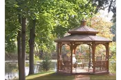 A wooden gazebo sits beside a pond surrounded by trees and leafy greenery.