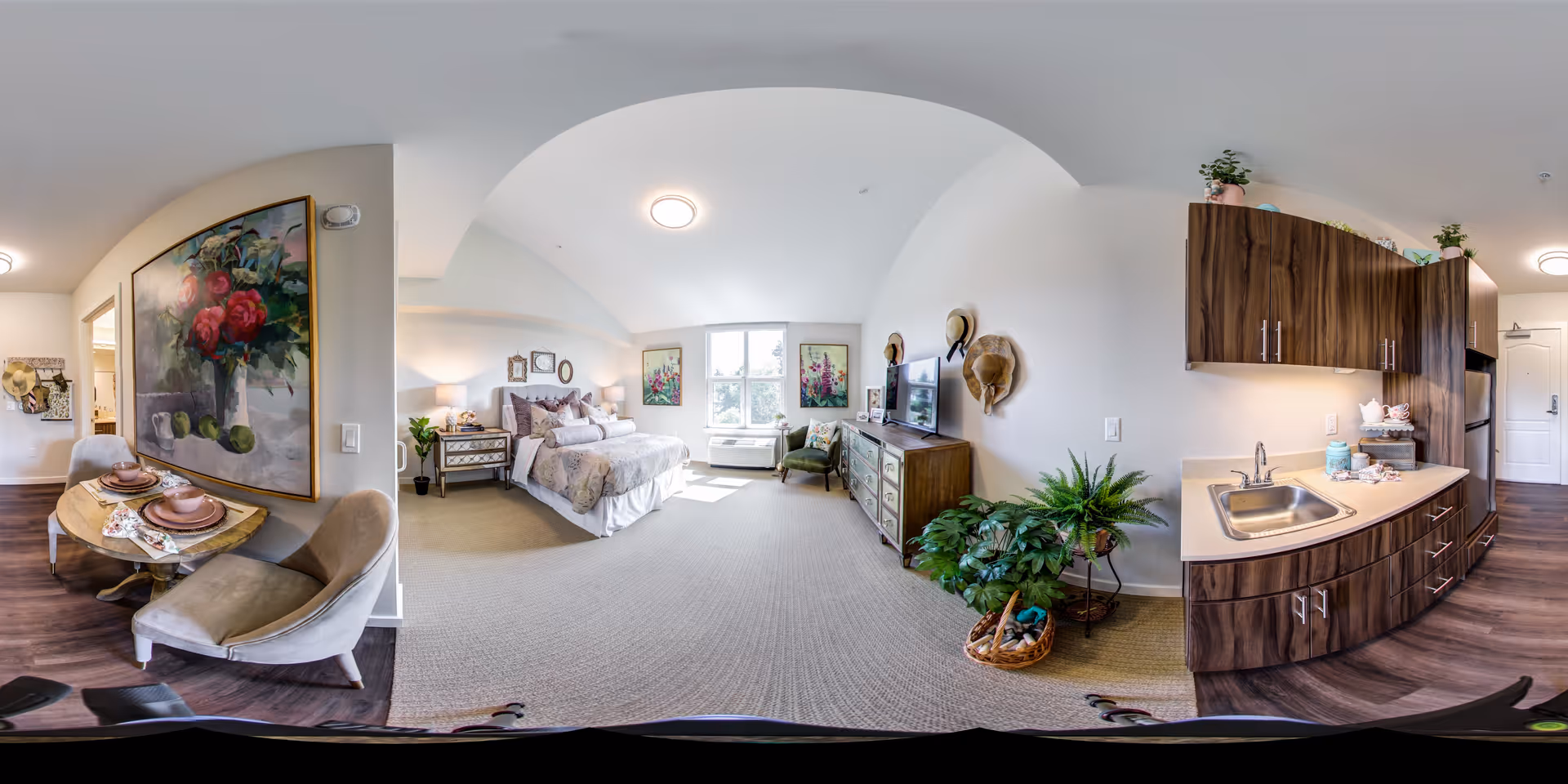 A spacious, well-lit senior living apartment featuring a cozy bedroom with a large bed, bedside tables with lamps, and floral artwork on the walls. Adjacent to the bedroom is a small dining area with a table set for two and cushioned chairs. The kitchenette has wooden cabinets, a sink, and a countertop with decorative items. The room is decorated with plants and hats hanging on the wall, creating a warm and inviting atmosphere.