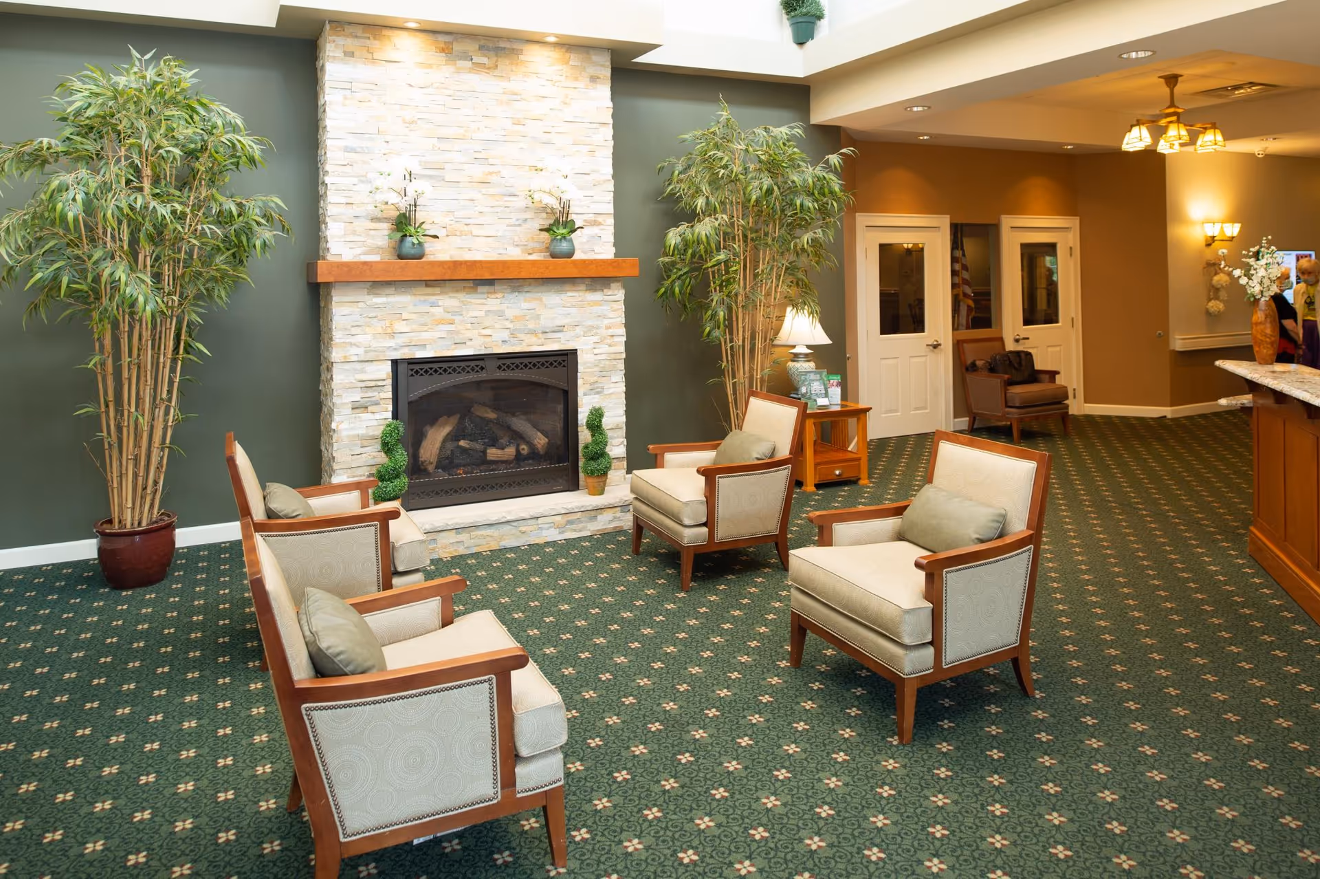 A cozy sitting area with four beige cushioned armchairs arranged around a stone fireplace with a wooden mantle. Two tall potted plants flank the fireplace. The room has green patterned carpet and warm lighting, with a small wooden side table holding a lamp and brochures. In the background, there are two white doors and a reception desk with flowers on the right side.