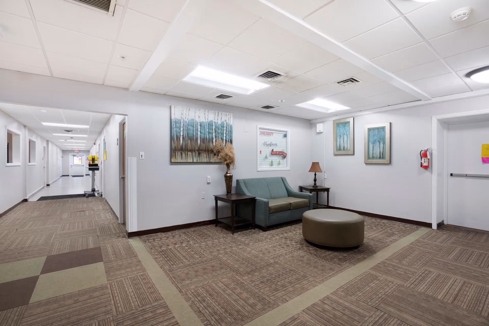 A well-lit senior living facility hallway with a seating area featuring a green loveseat, two side tables with a lamp and a vase, and a round ottoman. The walls are decorated with framed artwork and a holiday-themed window decoration. The floor is carpeted with a patterned design, and the ceiling has recessed lighting panels.