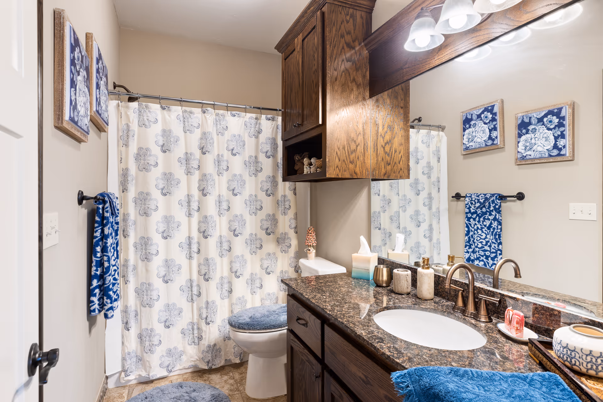 A bathroom with a granite countertop and an oval sink. Above the sink is a large mirror with three light fixtures. There are wooden cabinets above and below the countertop. A toilet with a blue fuzzy seat cover is next to a shower with a white curtain featuring a blue floral pattern. Blue towels hang on the wall, and two framed blue and white floral artworks are displayed above the towel rack. The floor is tiled.