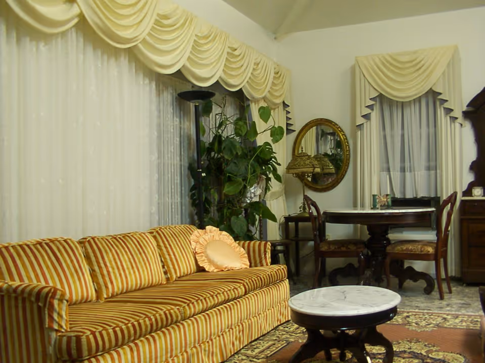 A cozy living room with a striped yellow and orange sofa adorned with a round peach pillow. The room features large windows with cream-colored draped curtains, a tall green plant, a round wooden table with two chairs, a small round marble-top coffee table, a decorative mirror on the wall, and a patterned rug on the floor.