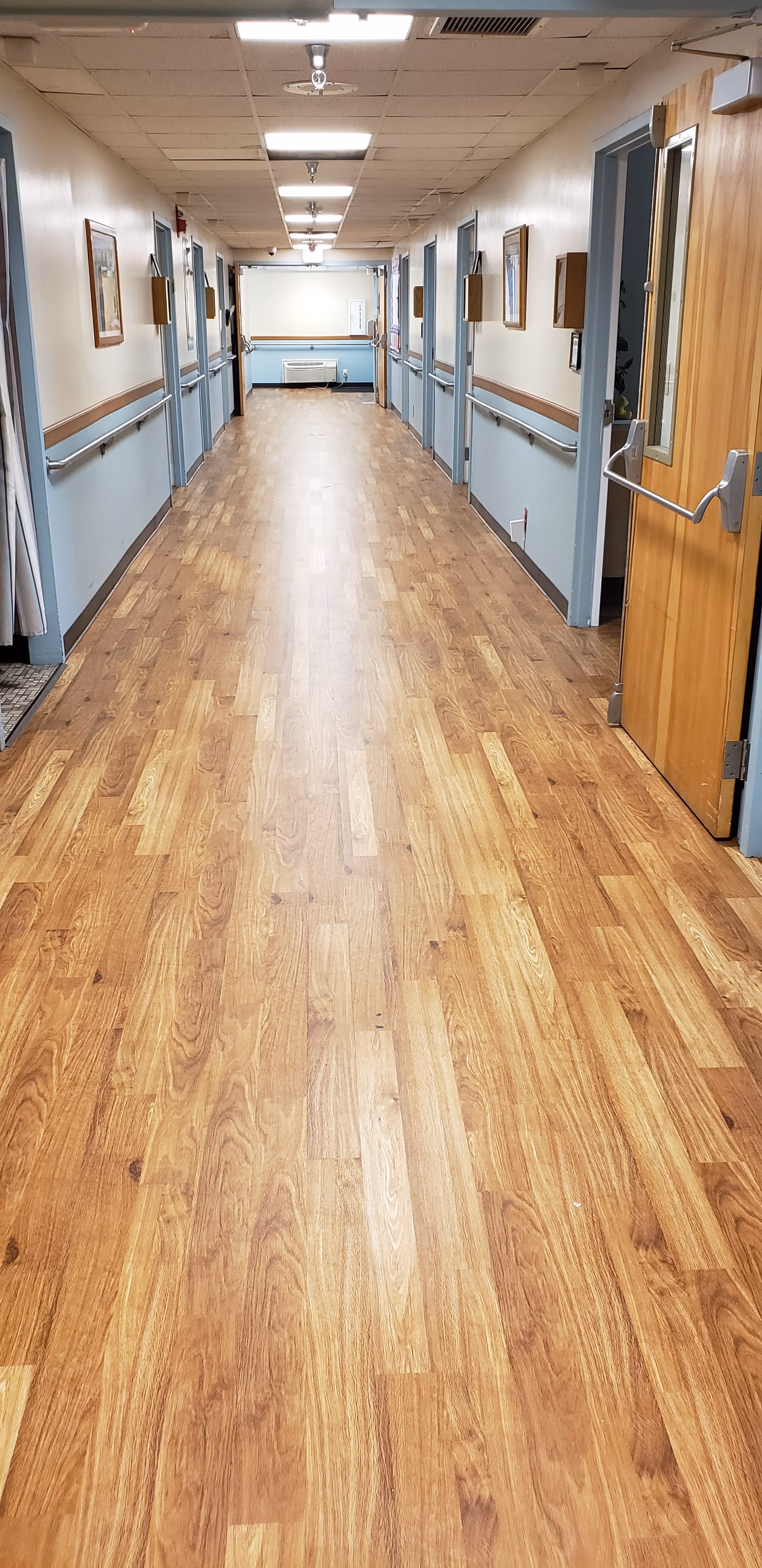 A long, empty hallway in a senior living facility with wood-patterned flooring, light blue lower walls with handrails, cream upper walls, and several closed doors on both sides. The ceiling has fluorescent lights and ventilation panels. At the end of the hallway, there is a whiteboard and a small bench.