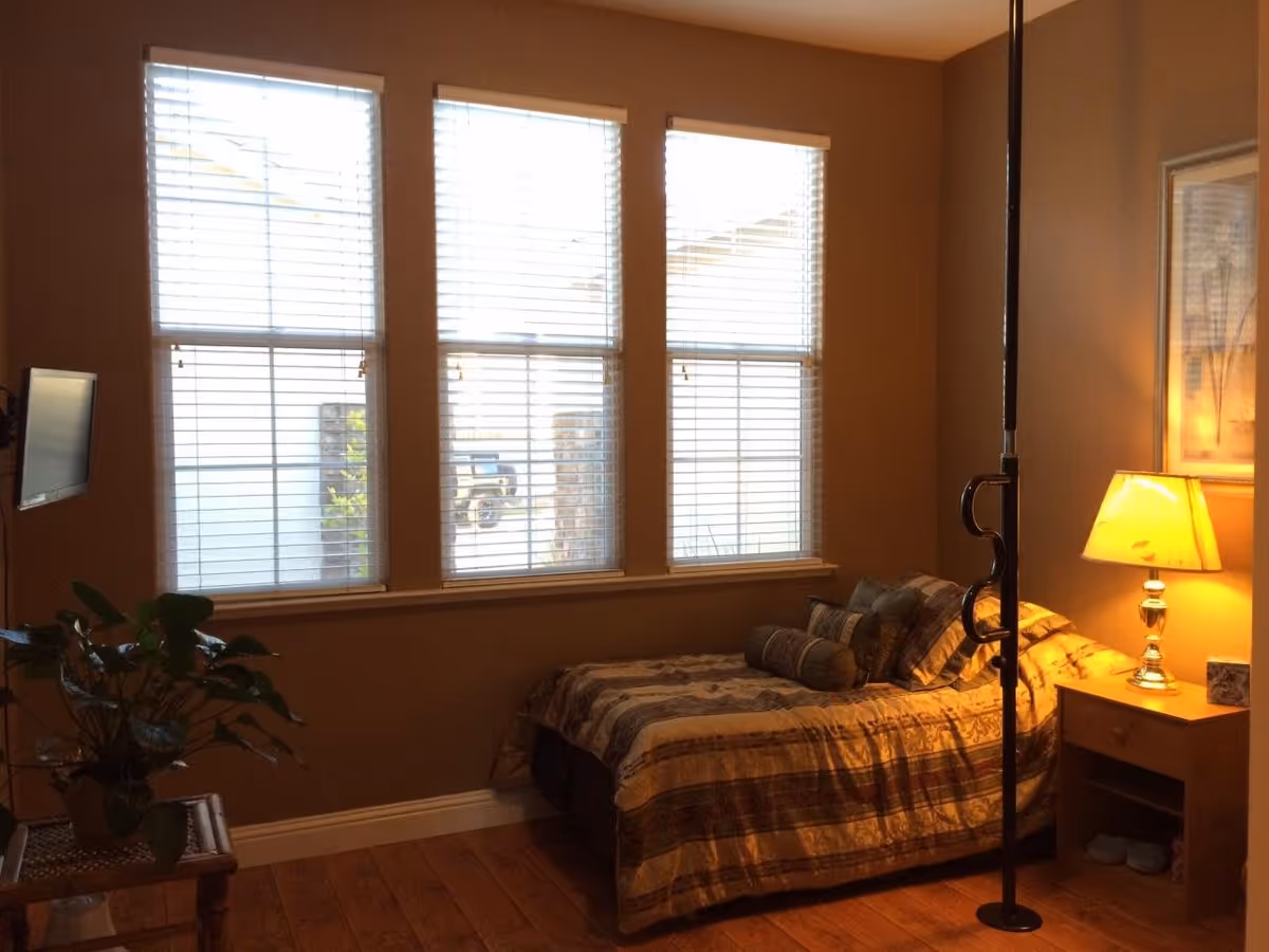 A cozy bedroom with a single bed covered in a patterned bedspread and several pillows. Next to the bed is a wooden nightstand with a lit lamp and a small box. Three large windows with blinds allow natural light into the room. A flat-screen TV is mounted on the wall to the left, and a green potted plant sits on a small table beneath it. The room has wooden flooring and warm-colored walls.